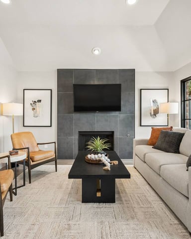 Modern living room with fireplace, beige sofa, tan leather chairs, black coffee table, and large windows overlooking trees